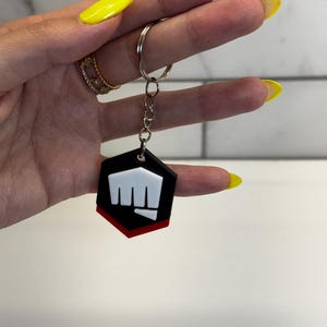 May include: A black and red keychain with a white fist logo. The keychain is hexagonal and attached to a silver keyring. The background is a white tile wall.