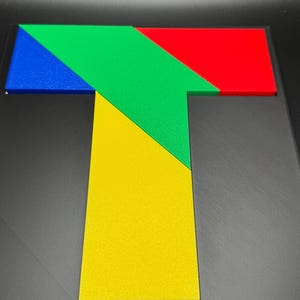 May include: A colorful wooden puzzle shaped like the letter T. The puzzle is made up of five pieces: a blue triangle, a green triangle, a red triangle, a yellow triangle, and a yellow square.