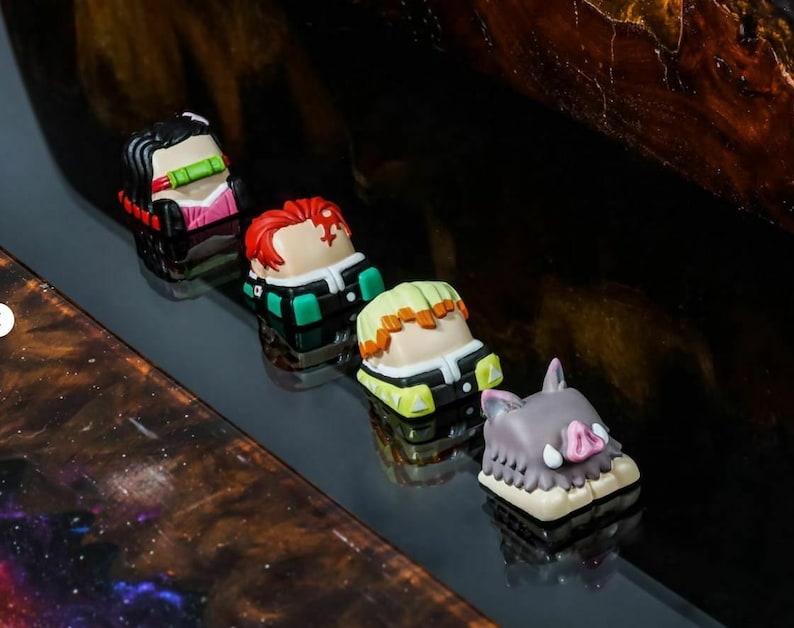 May include: Five colorful keycaps featuring characters from the anime Demon Slayer. The keycaps are made of plastic and have a 3D design. The characters include Tanjiro, Nezuko, Zenitsu, Inosuke, and the demon Muzan.