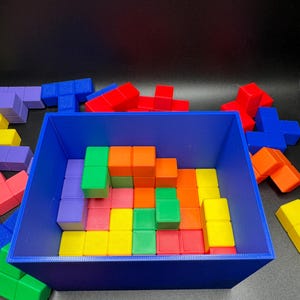May include: A blue rectangular container filled with colorful, geometric building blocks. The blocks are various shapes and colors, including red, green, orange, yellow, and purple. Some blocks are outside the container.