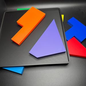 May include: A set of colorful geometric puzzle pieces on a black surface. The pieces are made of plastic and include a bright orange L-shaped piece, a purple triangle, a blue T-shaped piece, and a red rectangle.