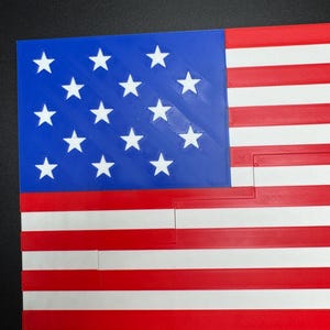 Puede incluir: Una bandera estadounidense impresa en 3D con un campo de estrellas azul y 13 rayas rojas y blancas.