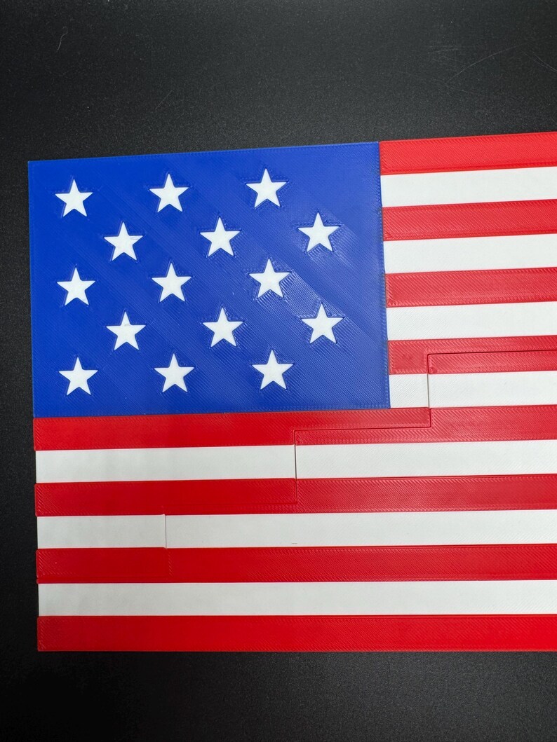 Puede incluir: Una bandera estadounidense impresa en 3D con un campo de estrellas azul y 13 rayas rojas y blancas.