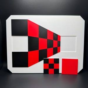 May include: A white rectangular art piece with a geometric design. The design features a red and black checkerboard pattern that transitions into a diagonal arrangement. Additional red and black squares are placed below the main design.