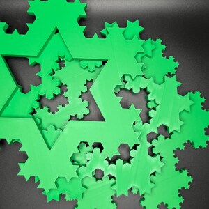 May include: A close-up of a green 3D printed fractal snowflake design. The snowflake is made up of many smaller snowflakes, creating a complex and intricate pattern.