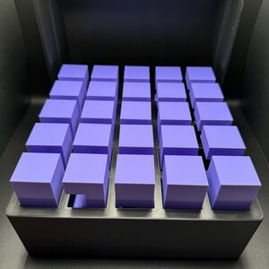 May include: A black box with a lid open, revealing a grid of 25 small, purple cubes.