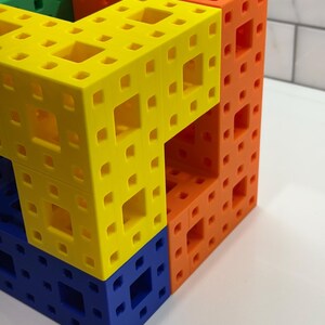 May include: A multi-colored cube made of interlocking plastic pieces. The cube is made of four different colors: green, yellow, orange, and blue. The cube is a three-dimensional puzzle.