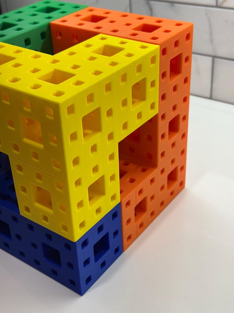May include: A multi-colored cube made of interlocking plastic pieces. The cube is made of four different colors: green, yellow, orange, and blue. The cube is a three-dimensional puzzle.
