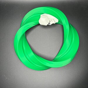 Może przedstawiać: Zielony plastikowy pierścień o skręconym wzorze. Pierścień wykonany jest z elastycznego materiału i ma gładką powierzchnię.