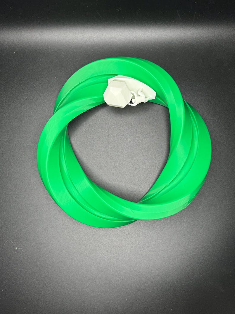 Może przedstawiać: Zielony plastikowy pierścień o skręconym wzorze. Pierścień wykonany jest z elastycznego materiału i ma gładką powierzchnię.