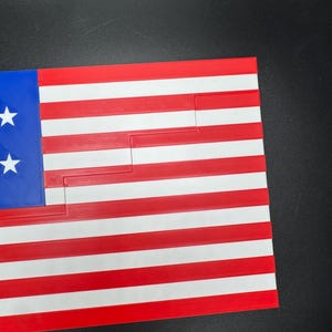 Puede incluir: Una bandera estadounidense roja, blanca y azul hecha de pl&aacute;stico. La bandera est&aacute; recortada de forma que crea un patr&oacute;n de escalera.