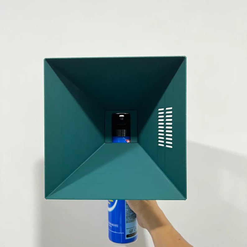 May include: A teal green spray bottle diffuser with a square opening and a series of small holes on the side. The diffuser is being held by a hand, and a blue and white spray bottle is being inserted into the opening.