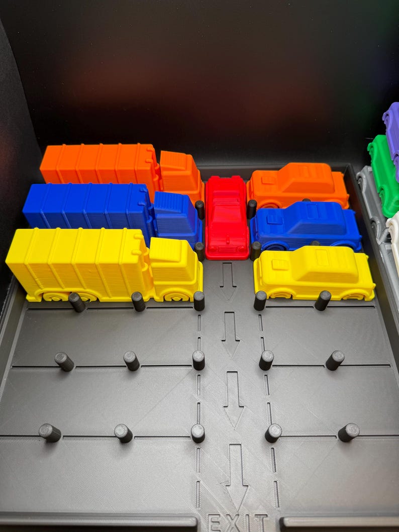 May include: A selection of brightly coloured toy lorries in shades of orange, blue, yellow, and red, arranged in a grey tray. The tray features a road design with arrows and the word "EXIT".