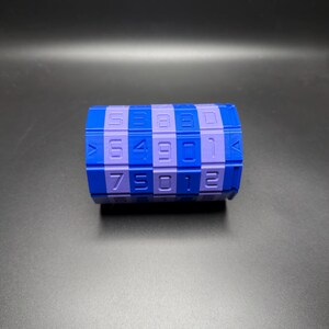 May include: A blue and purple combination lock toy with numbers. The toy is cylindrical with a checkerboard pattern of blue and purple squares. Numbers are printed on the purple squares. The background is a dark gray.