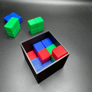 May include: A black square container filled with red, blue, and green cube-shaped puzzle pieces. Several loose cubes of the same colors are scattered around the container. The cubes are arranged in a pattern.