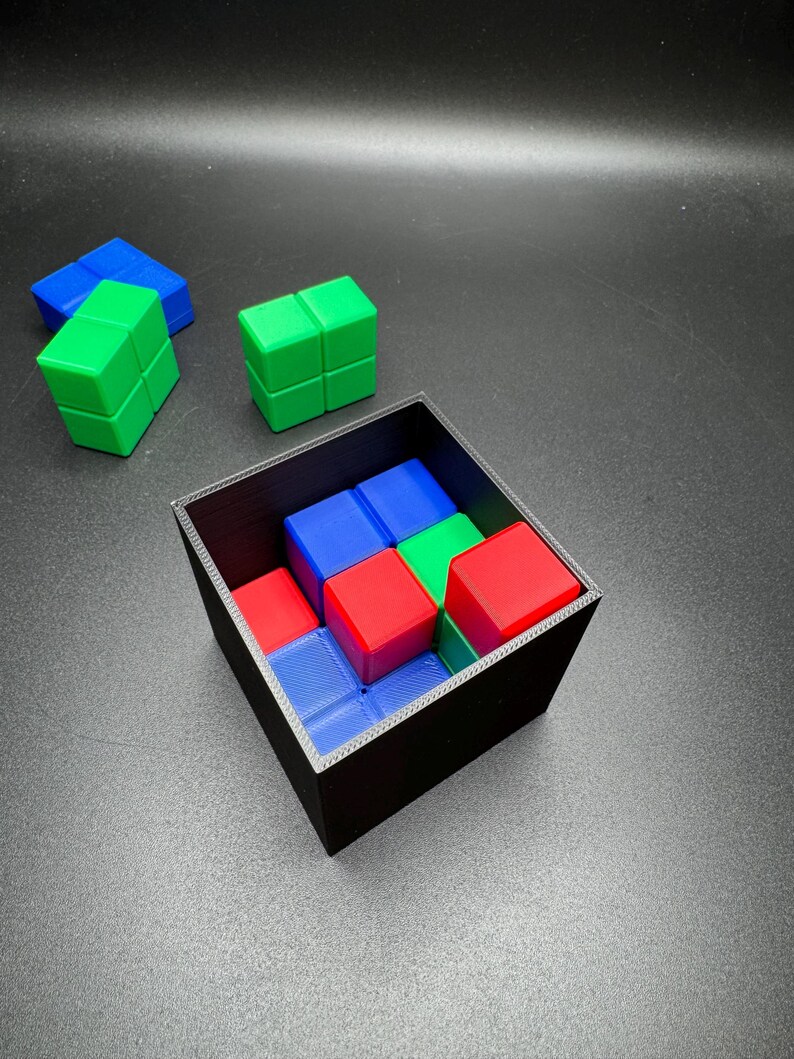 May include: A black square container filled with red, blue, and green cube-shaped puzzle pieces. Several loose cubes of the same colors are scattered around the container. The cubes are arranged in a pattern.