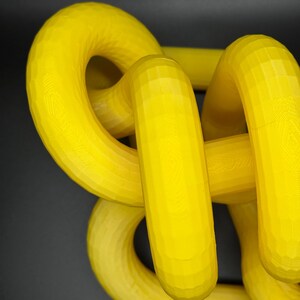 May include: A yellow 3D printed object with a complex, interwoven design. The object is made of multiple loops and curves, creating a visually interesting and abstract shape.