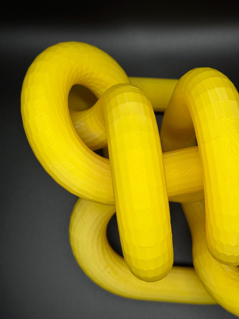 May include: A yellow 3D printed object with a complex, interwoven design. The object is made of multiple loops and curves, creating a visually interesting and abstract shape.
