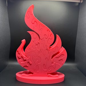 May include: A red flame-shaped puzzle, assembled and standing upright. The puzzle is made of interlocking pieces, creating the shape of a stylized flame. It sits on a circular base, all in a vibrant red color.