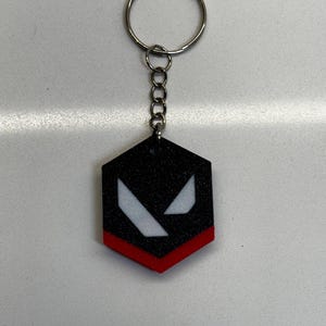 May include: Black and red keychain with a silver key ring. The keychain has a black hexagon shape with a red border and white angled shapes. A chain connects the hexagon to the key ring.