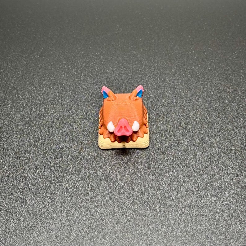 May include: A brown and beige keycap with a boar's head design. The boar has a pink snout and blue ears.
