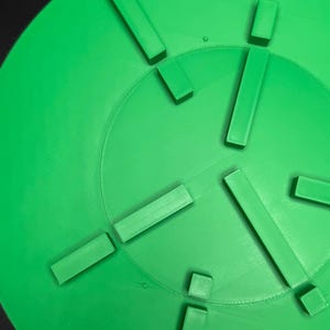 May include: Bright green, circular object with a series of rectangular and square protrusions. The object has a textured surface and a central circular indentation. The design suggests a functional or decorative item.