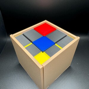 May include: A wooden box with a lid, containing nine small square blocks. The blocks are arranged in a 3x3 grid pattern. The blocks are coloured red, blue, yellow, and grey.