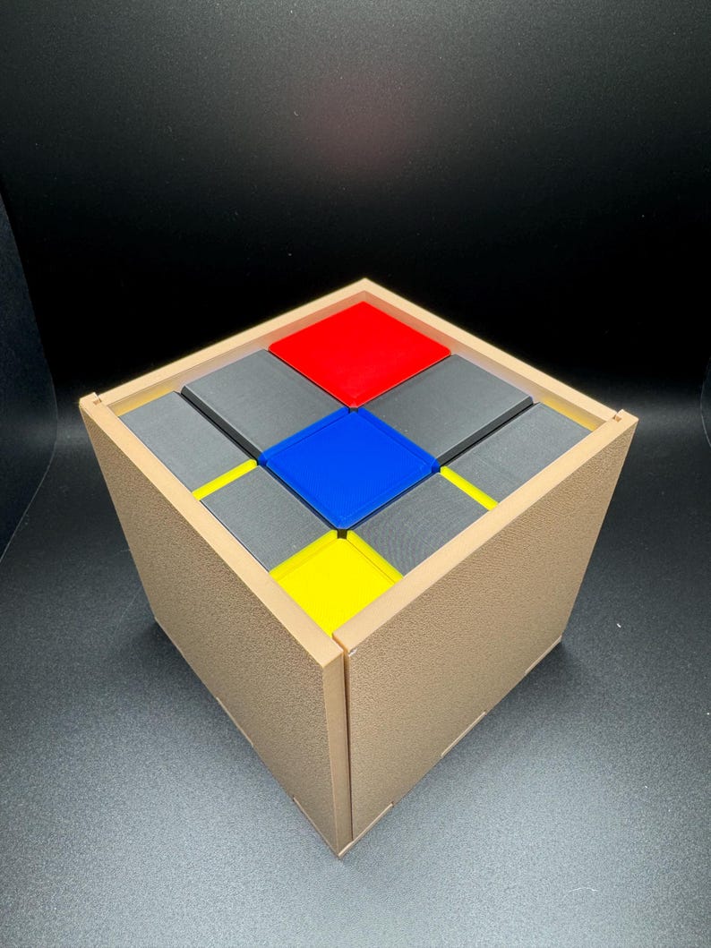 May include: A wooden box with a lid, containing nine small square blocks. The blocks are arranged in a 3x3 grid pattern. The blocks are coloured red, blue, yellow, and grey.