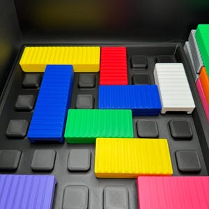 May include: A colorful puzzle game with various rectangular blocks in yellow, red, blue, green, white, purple, and pink. The blocks are arranged on a black tray with square indentations. The game appears to be a logic or spatial reasoning puzzle.