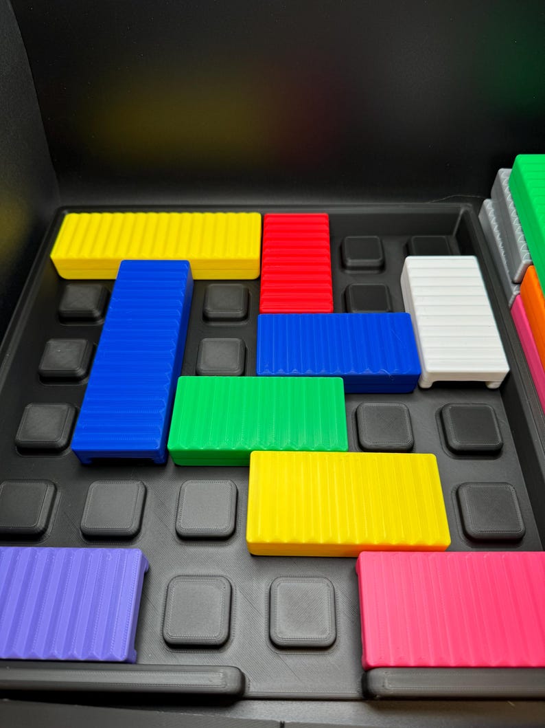 May include: A colorful puzzle game with various rectangular blocks in yellow, red, blue, green, white, purple, and pink. The blocks are arranged on a black tray with square indentations. The game appears to be a logic or spatial reasoning puzzle.