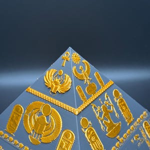 Puede incluir: Una pir&aacute;mide gris con jerogl&iacute;ficos y s&iacute;mbolos egipcios dorados. La superficie de la pir&aacute;mide est&aacute; detallada con varias figuras y patrones en relieve, mostrando el arte y el dise&ntilde;o del antiguo Egipto.