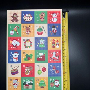 May include: A Christmas advent calendar with 24 colorful squares, each featuring a festive illustration and a number. The calendar is approximately 9 inches tall. The illustrations include a gingerbread man, snowman, and Santa Claus.