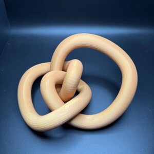 May include: A wooden sculpture of a knot in a light brown color. The knot is made of three intertwined loops.