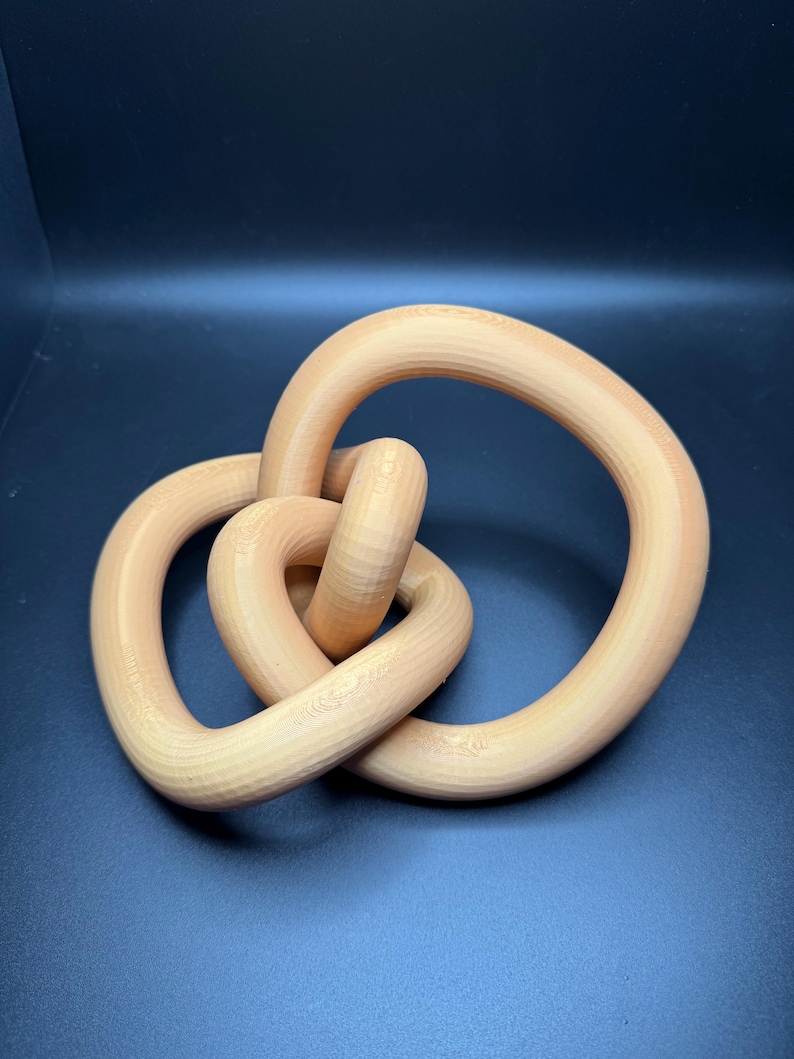 May include: A wooden sculpture of a knot in a light brown color. The knot is made of three intertwined loops.