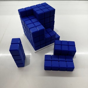 May include: A blue 3D puzzle made of small cubes. The puzzle is disassembled into four pieces. The largest piece is a cube with a missing corner. The other pieces are a long rectangular prism, a short rectangular prism, and a small cube.
