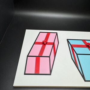 May include: Two 3D-printed gift boxes on a white surface. One is pink with red ribbon and a bow, the other is blue with red ribbon and a bow. Both have black outlines.
