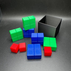 Könnte beinhalten: Eine Ansammlung farbiger Puzzelwürfel in Rot, Grün und Blau, zusammen mit einem schwarzen quadratischen Behälter. Die Würfel sind in einem verstreuten Muster angeordnet, was auf ein Spiel oder ein Rätsel hindeutet. Das Bild hat einen dunklen Hintergrund.