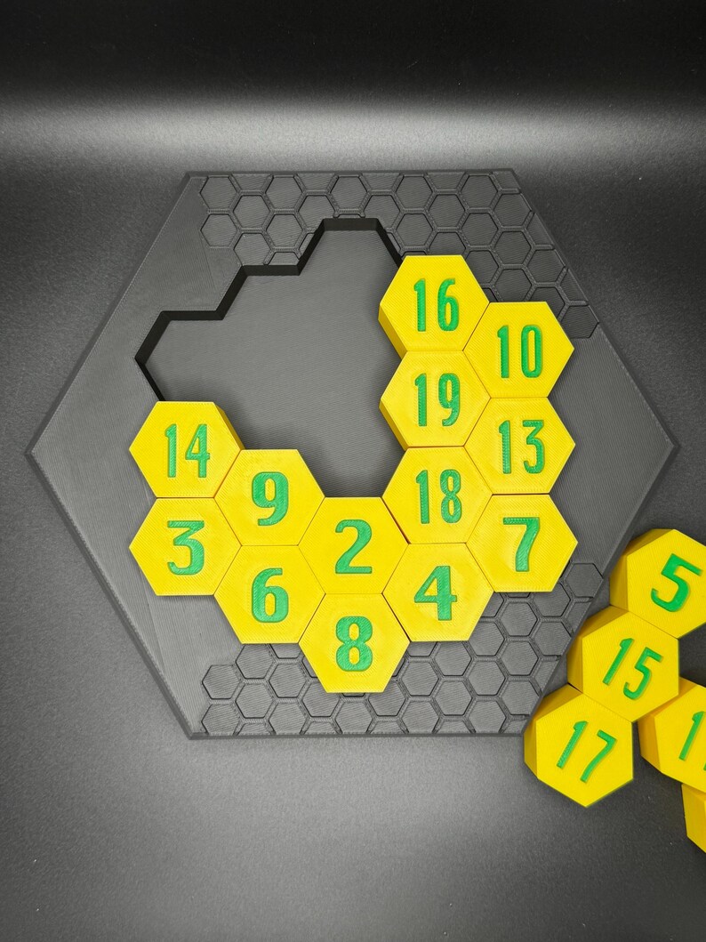 Puede incluir: Un juego de rompecabezas hexagonal con fichas hexagonales amarillas numeradas del 1 al 19. Las fichas est&aacute;n dispuestas en un patr&oacute;n de panal, con algunas fichas que faltan. El rompecabezas est&aacute; hecho de pl&aacute;stico negro.