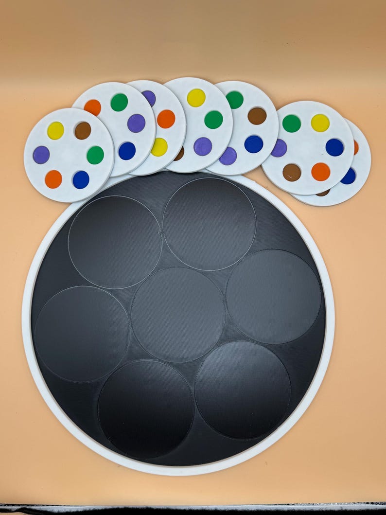 May include: A circular game board with a white border and a black interior, featuring seven black circles. Above, there are seven white circular pieces, each with eight colored dots: orange, yellow, green, purple, blue, brown.