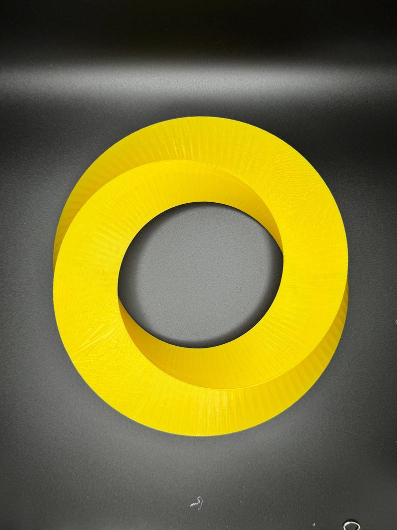May include: A bright yellow, 3D printed, Mobius strip shaped object. The object is a continuous loop with a hole in the center.