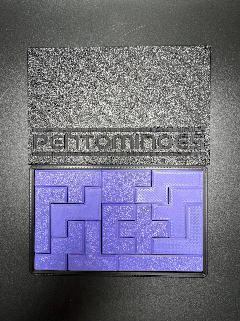 May include: A purple plastic pentominoes puzzle set in a black plastic case with the word "PENTOMINOES" printed on the lid.