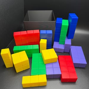May include: A collection of colorful, geometric building blocks in various shapes and sizes. The blocks are in shades of red, yellow, green, blue, and purple. A dark brown box is in the background. The blocks are arranged on a dark surface.