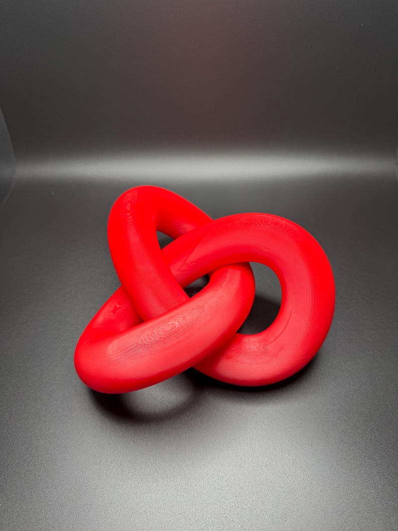 May include: A red, three-dimensional knot sculpture. The knot is made of smooth, rounded plastic and is tied in a complex pattern.