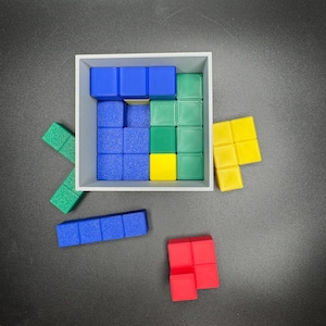 Puede incluir: Un conjunto de bloques de madera de colores en varias formas y tama&ntilde;os. Los bloques est&aacute;n dispuestos en una caja cuadrada, con algunos bloques fuera de la caja. Los bloques son azules, verdes, amarillos y rojos.
