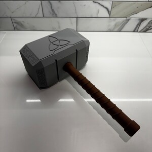 Peut inclure: Un marteau en plastique gris jouet avec une poign&eacute;e brune. Le marteau a un motif en relief d'un symbole triquetra sur la t&ecirc;te.