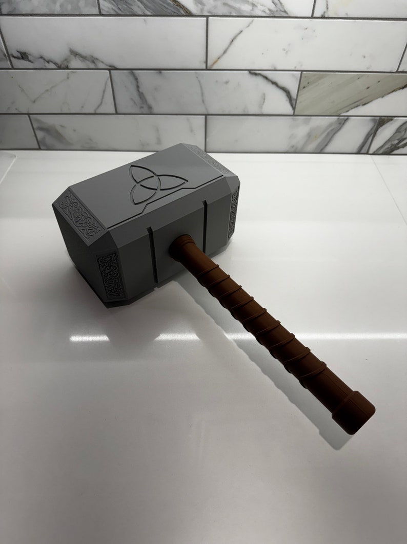 Peut inclure: Un marteau en plastique gris jouet avec une poign&eacute;e brune. Le marteau a un motif en relief d'un symbole triquetra sur la t&ecirc;te.