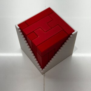 Puede incluir: Un rompecabezas 3D rojo en forma de cubo, ensamblado y colocado en un marco de pl&aacute;stico blanco.