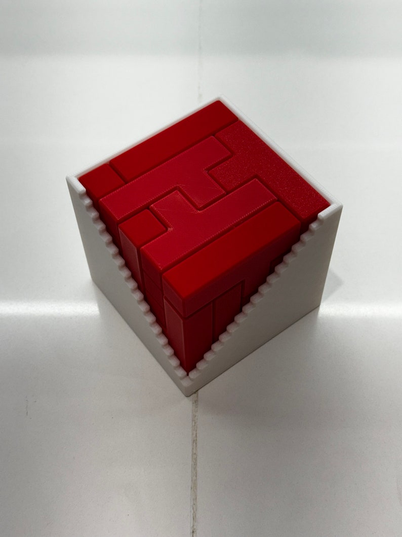 Puede incluir: Un rompecabezas 3D rojo en forma de cubo, ensamblado y colocado en un marco de pl&aacute;stico blanco.