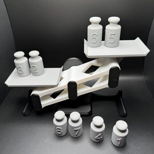 May include: A white plastic balance scale with six gray plastic weights. The weights are numbered 1, 2, 3, 4, 5, 6, and 7. The scale is tipped to the left side.