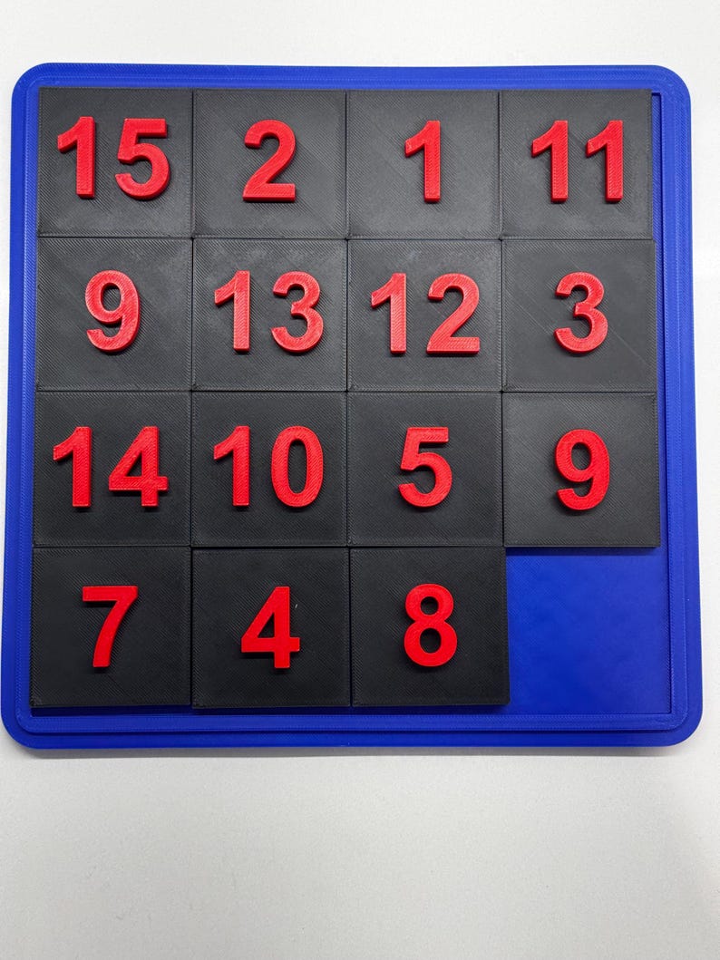 K&ouml;nnte beinhalten: Ein schwarzes und blaues Puzzlespiel mit roten Zahlen von 1 bis 15. Die Zahlen sind in einem 4x4-Raster angeordnet, wobei ein Quadrat fehlt.
